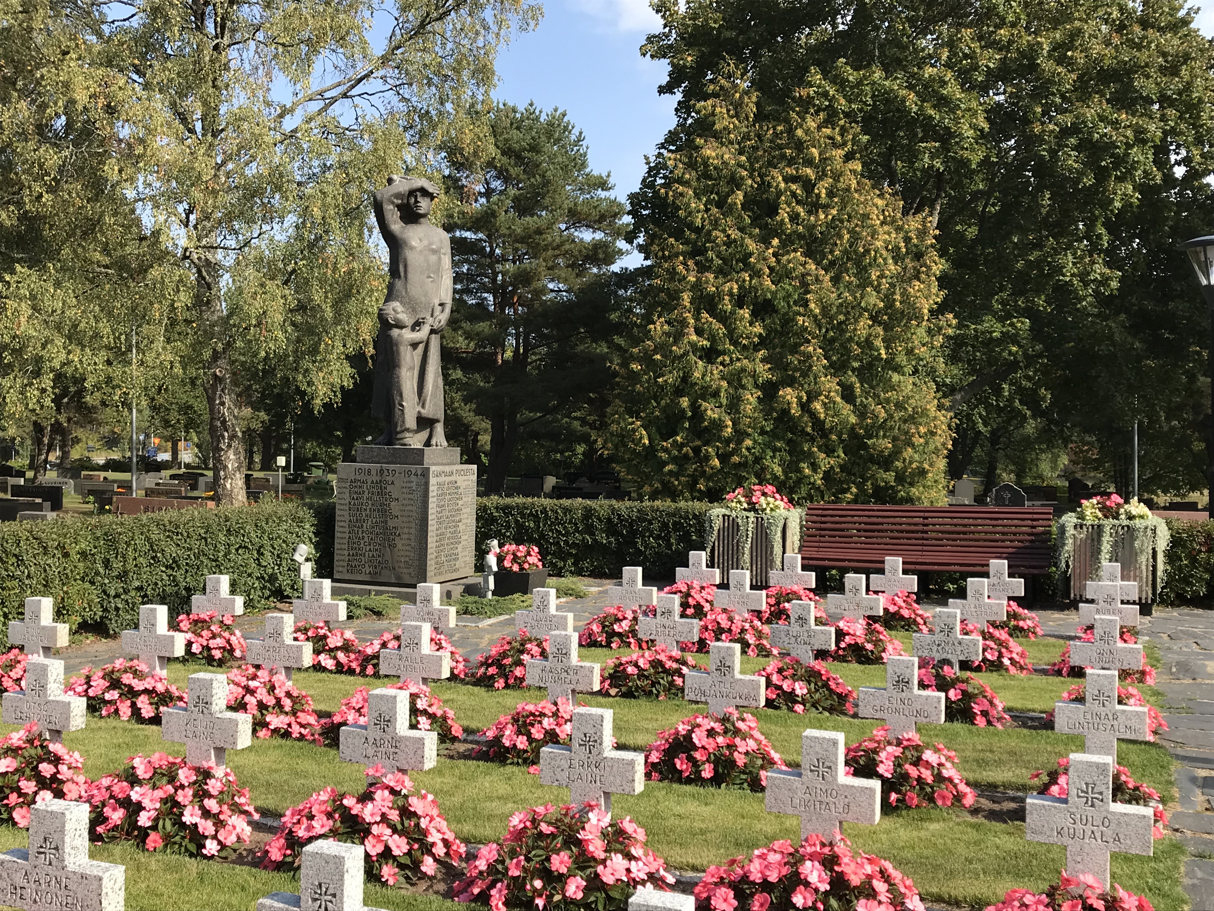 Sankarihaudan muistomerkki, jossa äiti tähyilee merelle pitäen lapsen kädestä kiinni. Vaaleat, kiviset kaatuneiden hautaristit, joiden edessä vaaleanpunaiset kukat.