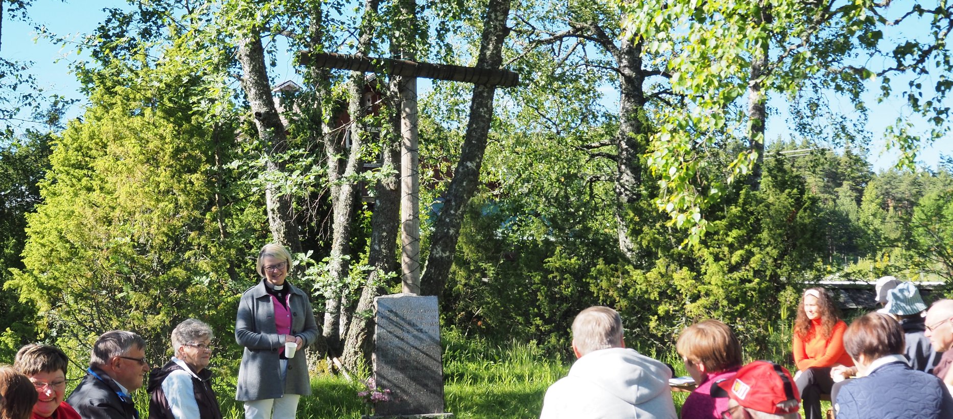 Vihreitä pensaita ja puita kivisen muistomerkin ja ristin takana. Pappi ja seurakuntalaisia juomassa kahvia ennen hartautta.